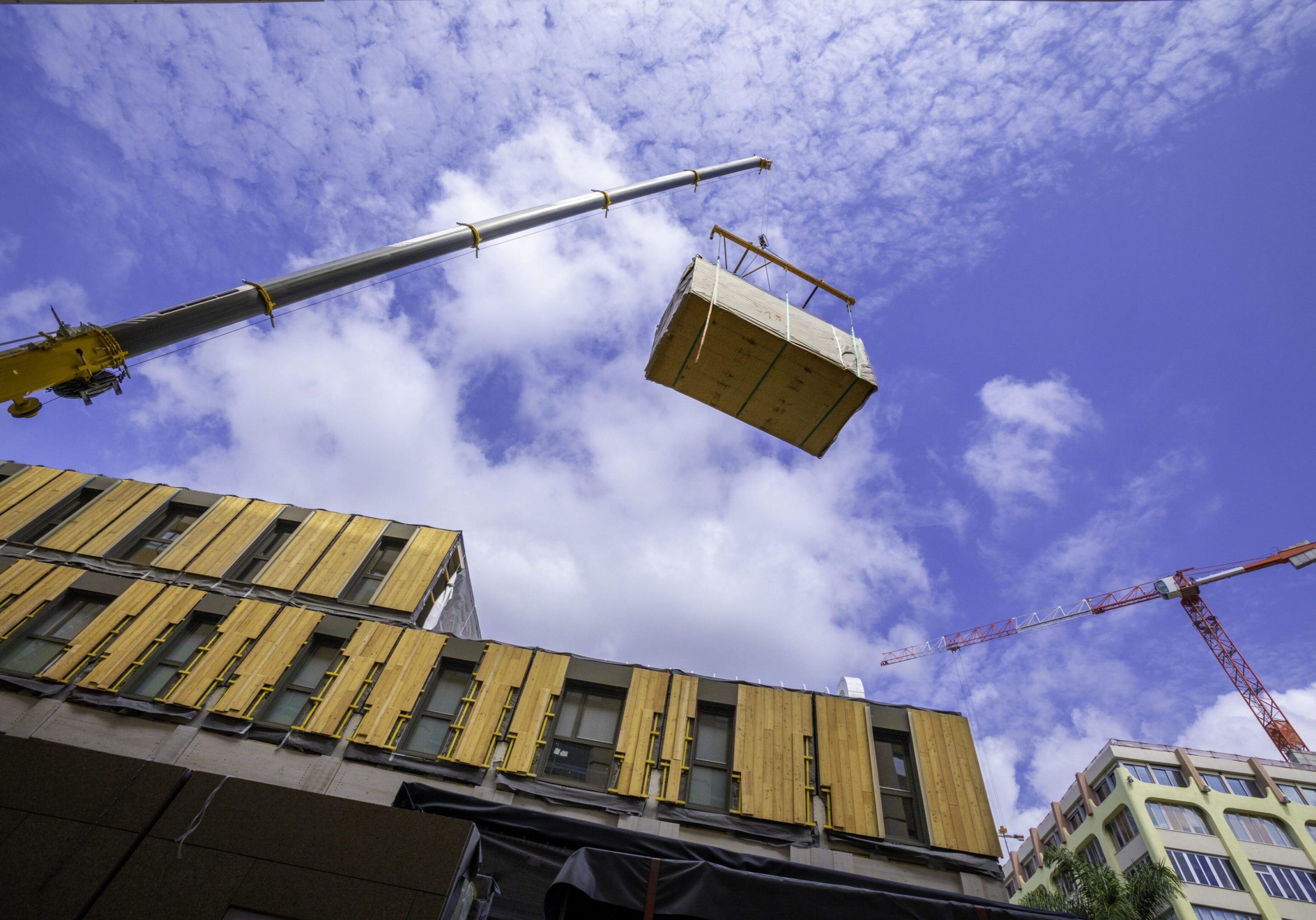 Pre-fabricated modular wood commercial building being assembled with crane A crane lifts a large rectangular container against a partly cloudy sky. Buildings with striped facades and another crane are seen in the background.